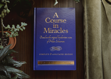 Load image into Gallery viewer, Front cover of A Course in Miracles Complete and Annotated Edition hardcover book, displayed flat on a dark wooden surface surrounded by green indoor plants and curtains.